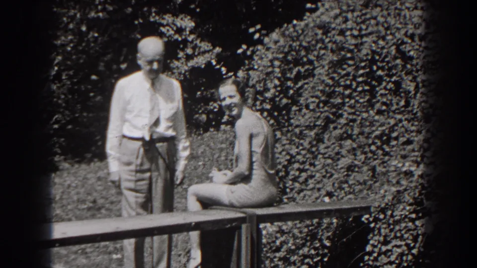 A young woman in a swimsuit sits on a wooden bench as an older man in a light shirt and tie stands beside her in the garden.