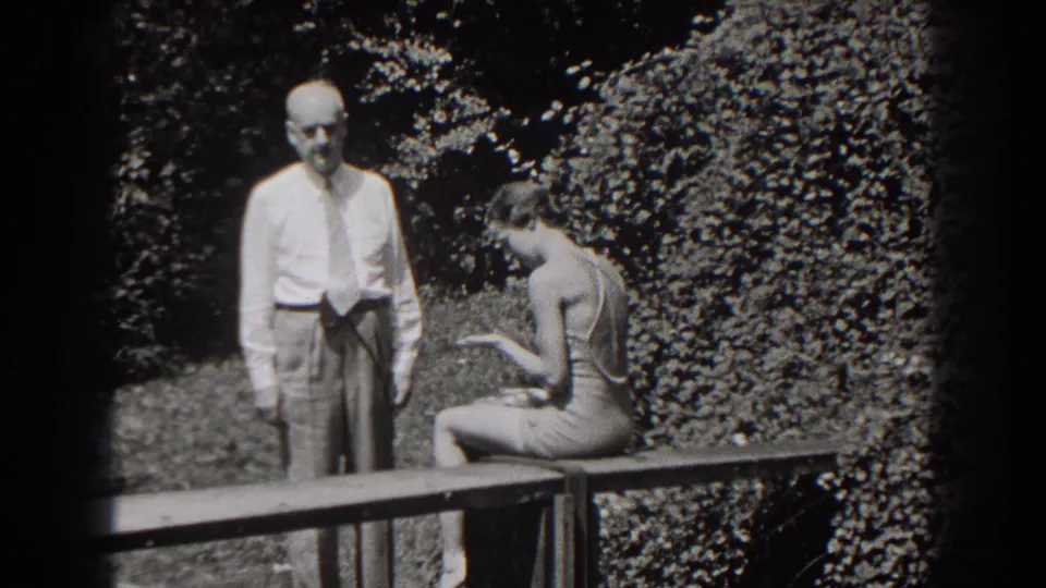 The seated young woman looks down at a small object or hat while the older man stands beside her in 1938 New Jersey garden footage.