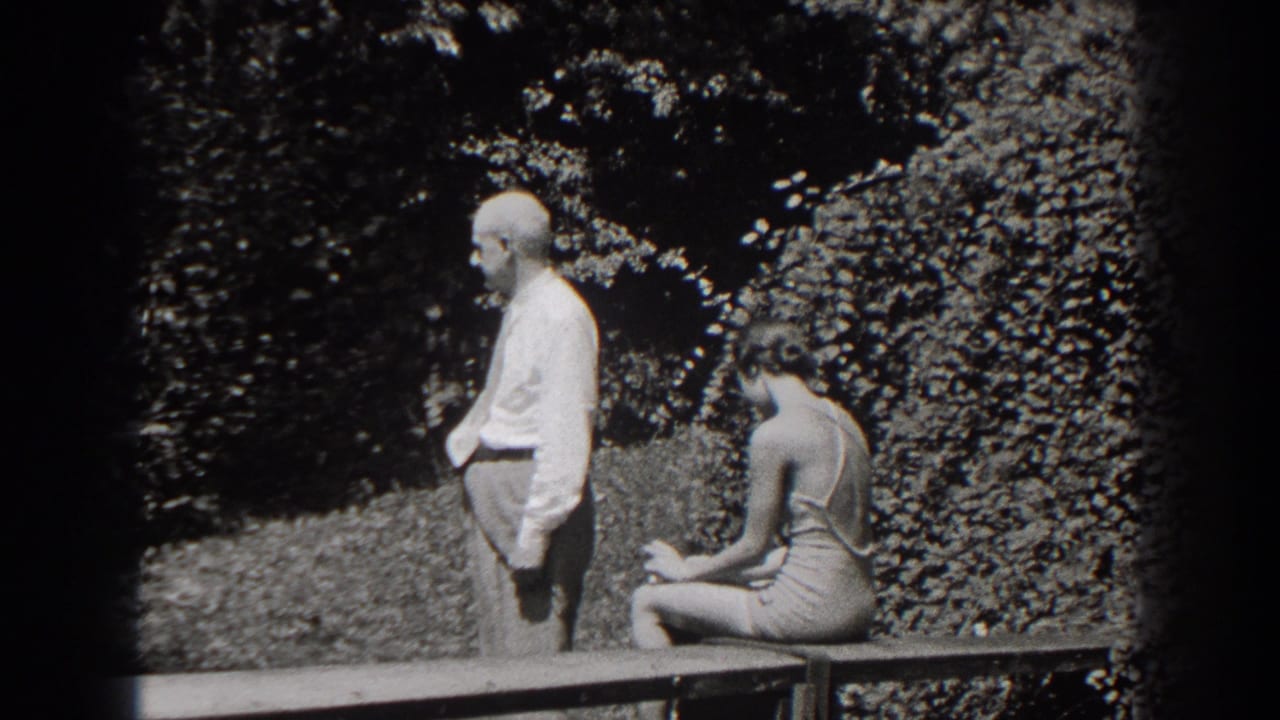 Older man standing beside a seated young woman on a garden bench.
