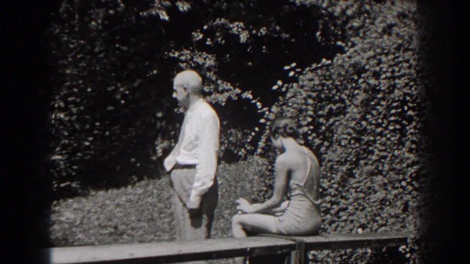 In a 1938 New Jersey garden scene, an older man stands with a cane beside a young woman seated on a wooden bench.
