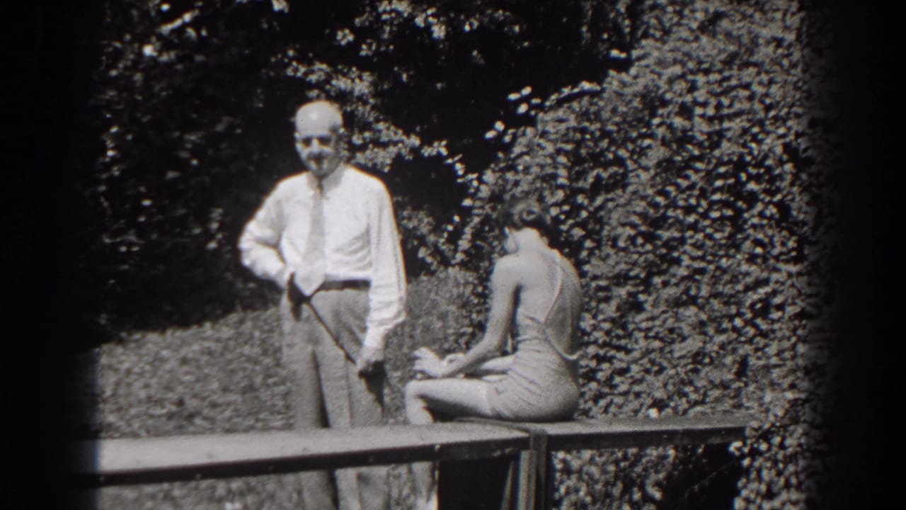 The man faces toward the camera with a cane at his side as the young woman sits turned away on the garden bench.