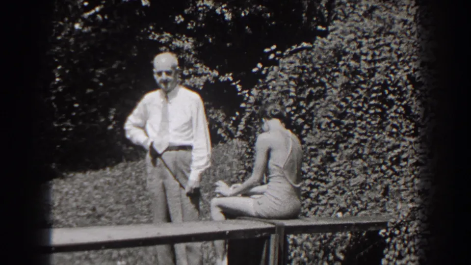 The man faces toward the camera with a cane at his side as the young woman sits turned away on the garden bench.