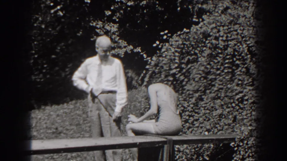 The garden home-movie frame shows the man standing with one hand at his waist while the seated young woman bends forward.