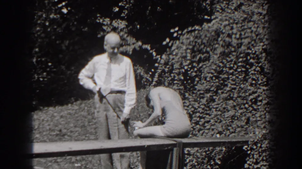 In 1938 New Jersey home-movie footage, a man in a white shirt and tie stands near a woman in a light swimsuit on a garden bench.