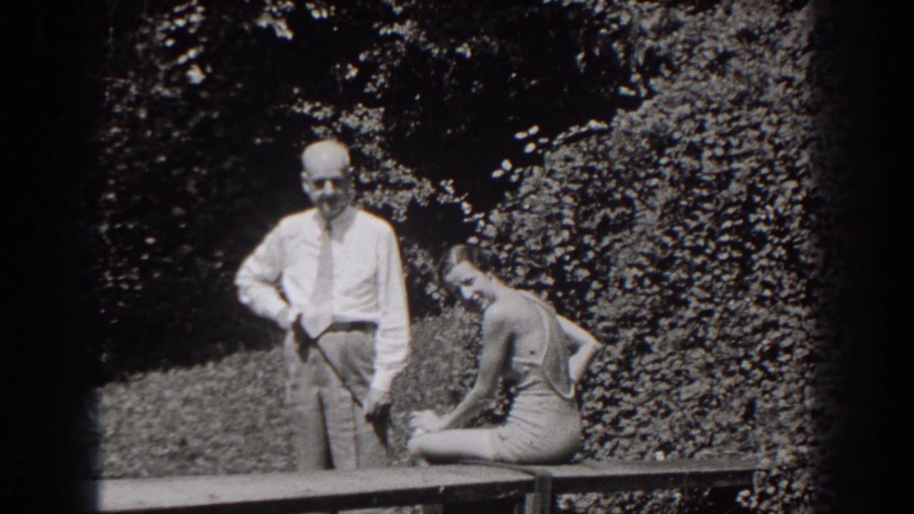 The woman turns and smiles toward the camera as the man stands next to her on a wooden bench in the 1938 New Jersey garden scene.