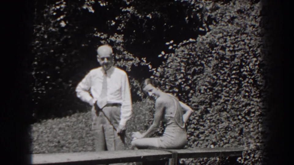 The woman turns and smiles toward the camera as the man stands next to her on a wooden bench in the 1938 New Jersey garden scene.