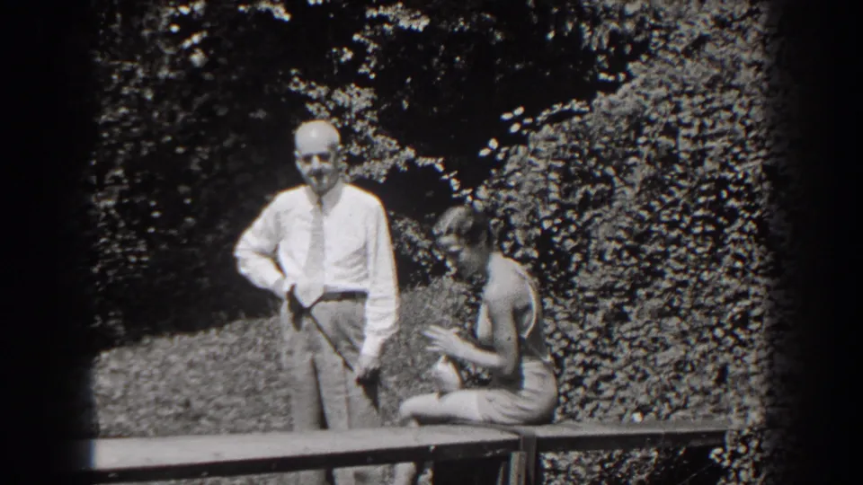 A man in a shirt and tie stands by the bench while the seated woman looks down, with foliage and dappled light behind them.