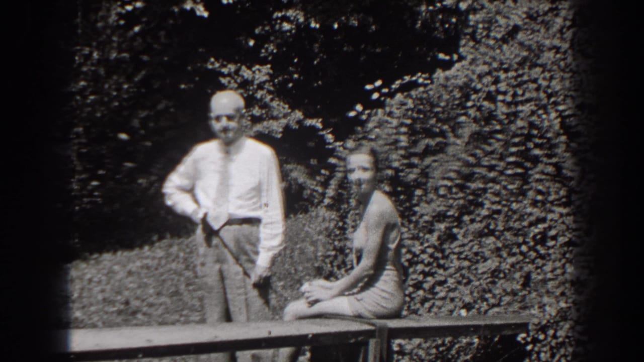 In 1938 New Jersey home-movie footage, a man in a shirt and tie stands beside a swimsuit-clad woman seated on a garden bench.
