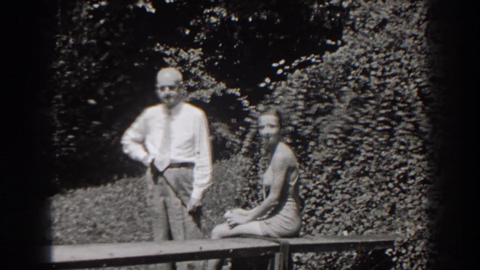 In 1938 New Jersey home-movie footage, a man in a shirt and tie stands beside a swimsuit-clad woman seated on a garden bench.