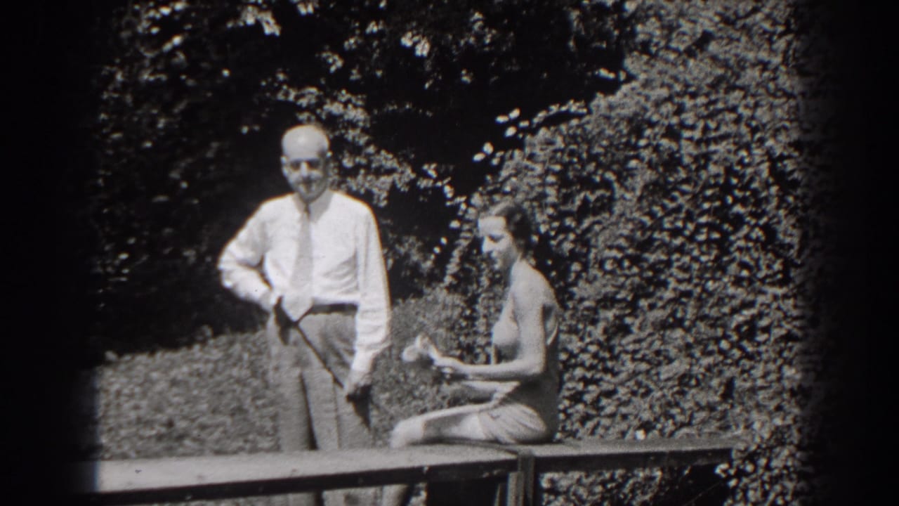 The woman seated on the garden bench appears to hold or adjust a small item as the man stands beside her in 1938 New Jersey footage.