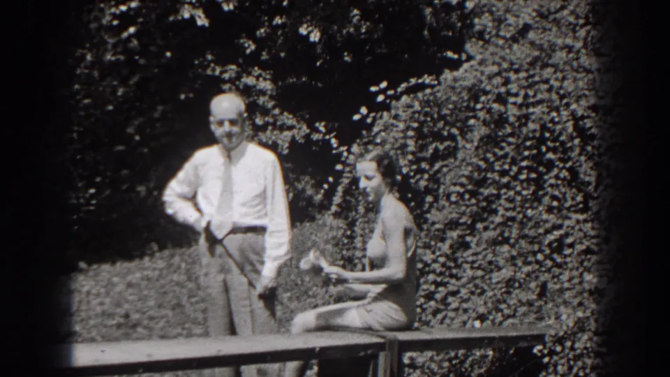 The woman seated on the garden bench appears to hold or adjust a small item as the man stands beside her in 1938 New Jersey footage.