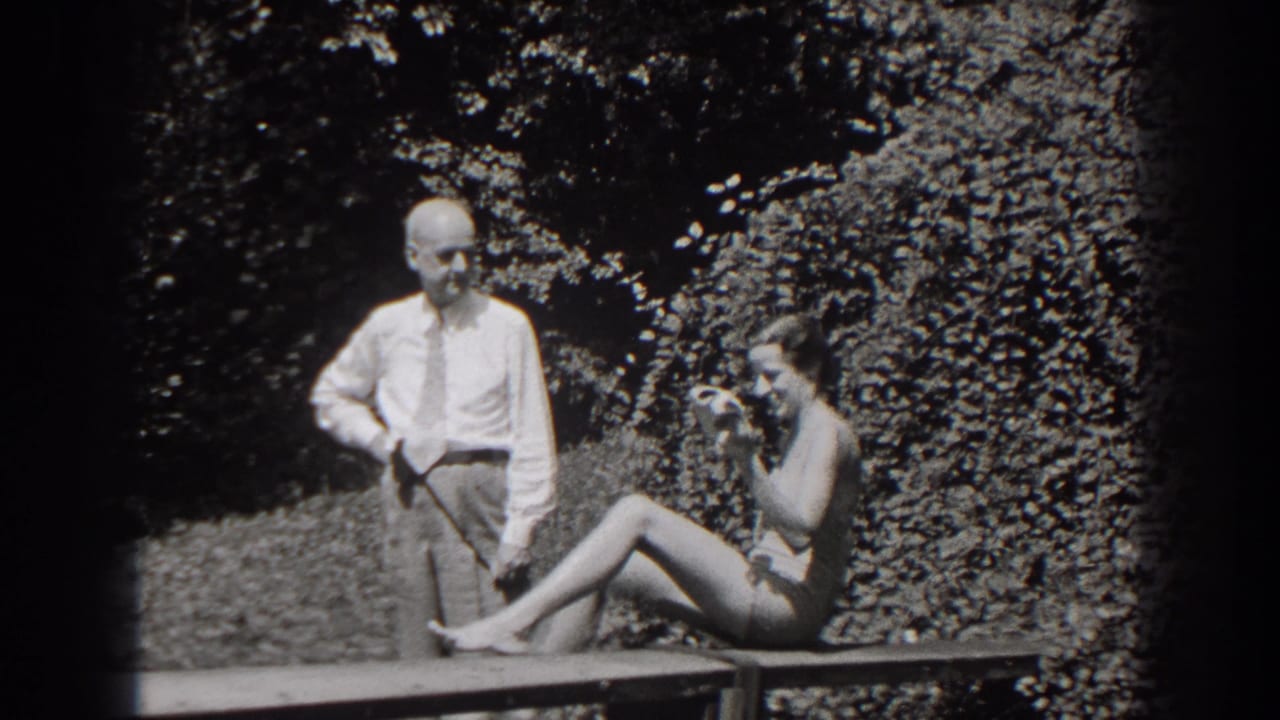 The woman shifts sideways on the bench, raising a hand near her face, while the man stands in the same garden pose.