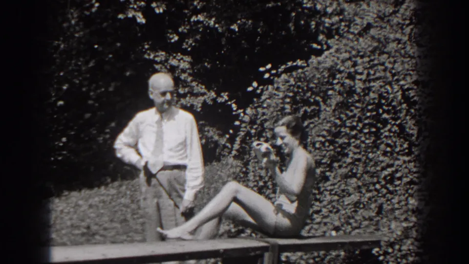 The woman shifts sideways on the bench, raising a hand near her face, while the man stands in the same garden pose.