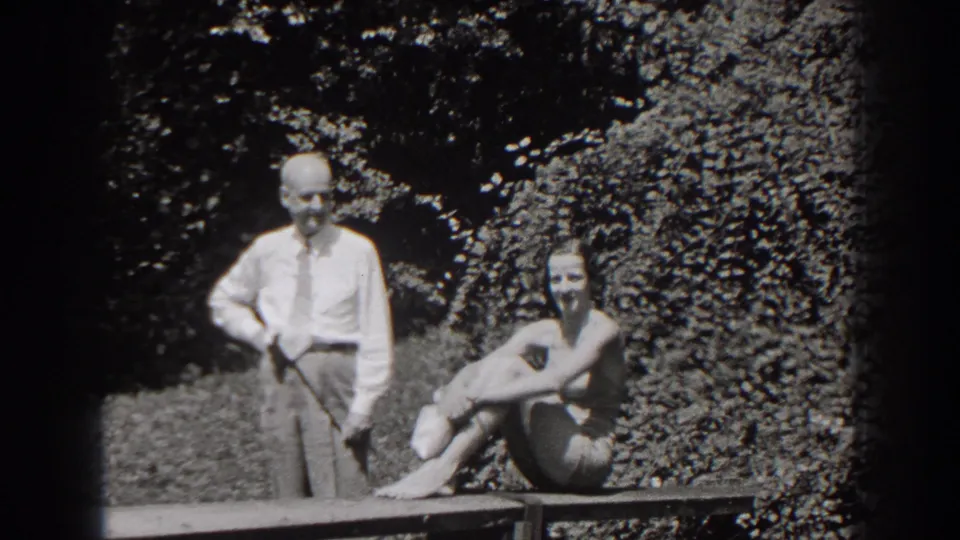 1938 New Jersey home-movie frame of a man holding a golf club near a seated woman in a bathing suit by leafy hedges.