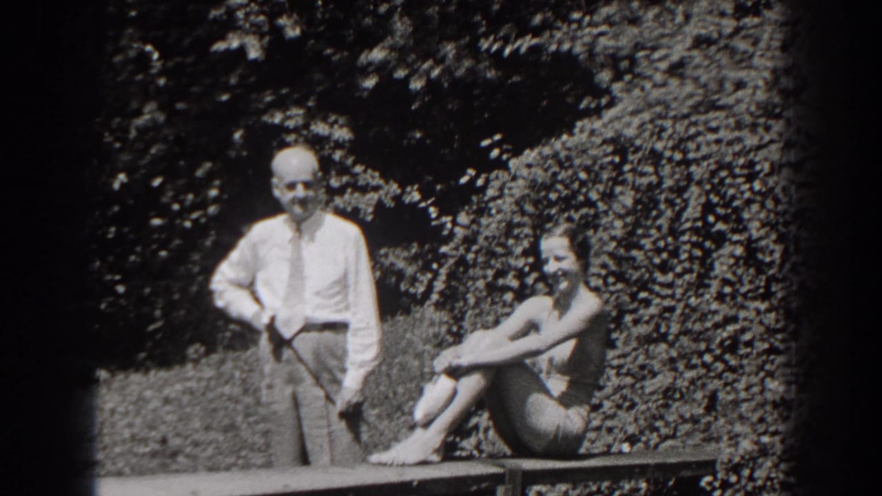 Standing man and seated woman smile outdoors against dense foliage.