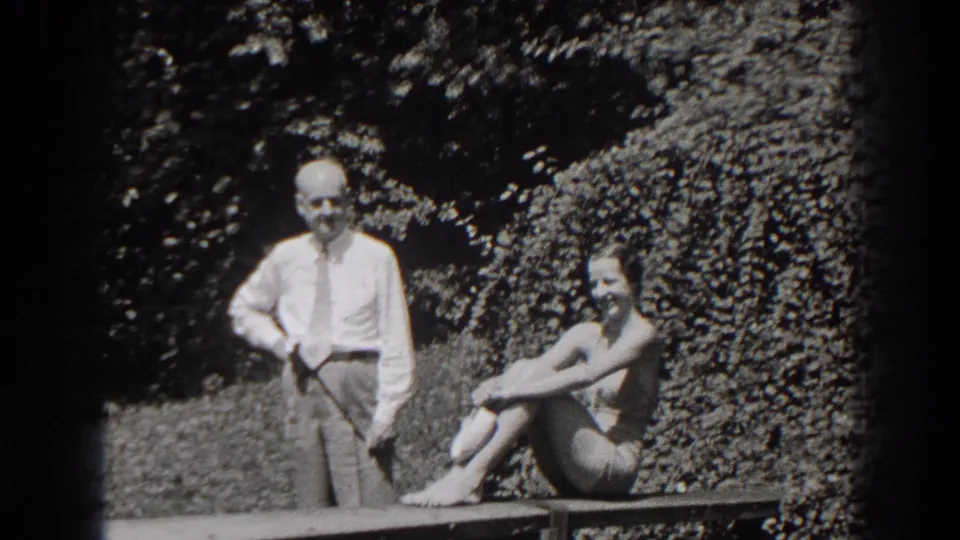 1938 New Jersey frame showing a standing man and seated woman in a bathing suit smiling in a sunlit garden.