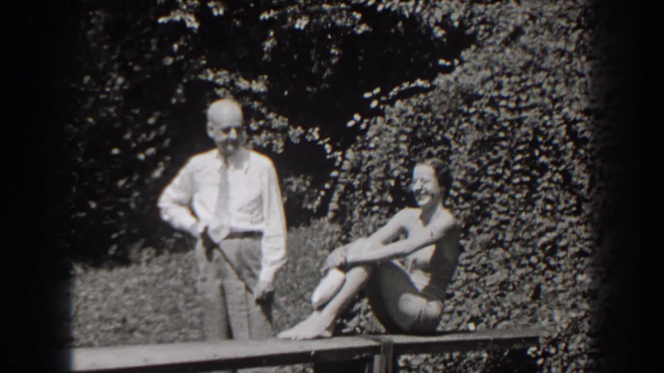 1938 New Jersey home-movie frame of a smiling seated woman in a bathing suit with a man standing beside her outdoors.
