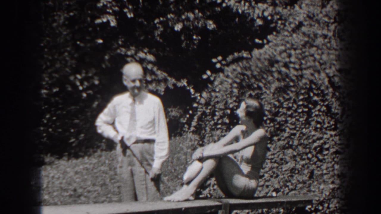 Older man stands beside a bench while a woman sits outdoors near dense foliage.