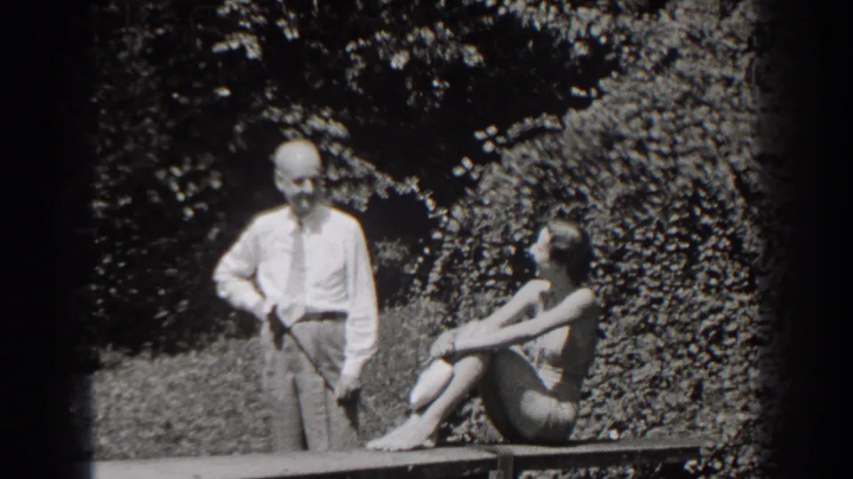 In a 1938 New Jersey home-movie frame, an older man stands by a bench as an adult woman sits and looks toward him in a garden.