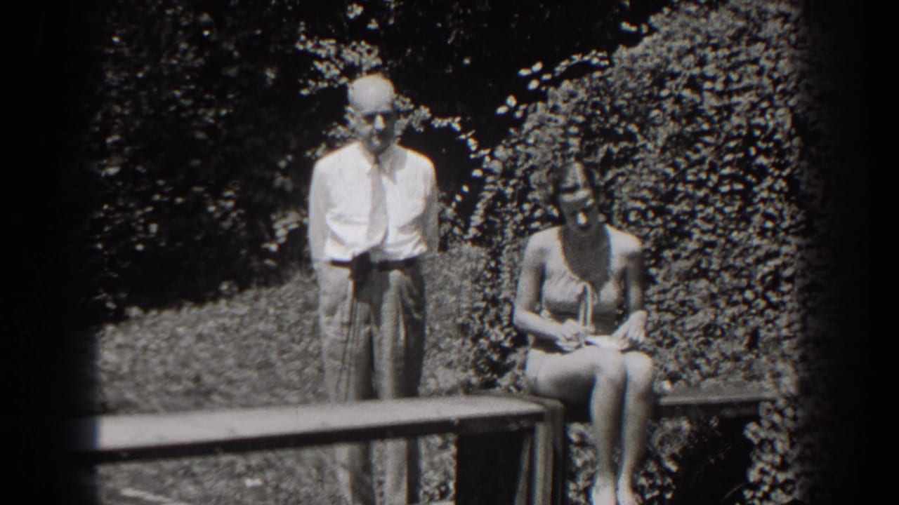 Man stands behind a bench while the seated woman looks down at something in her hands.