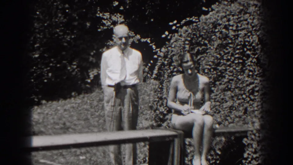 The man stands near the bench in the garden while the seated woman in a sleeveless outfit looks downward, possibly at her hands.
