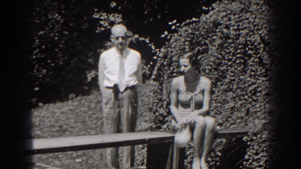 A 1938 New Jersey frame shows the woman seated on the bench and the older man standing behind the bench rail against dense foliage.