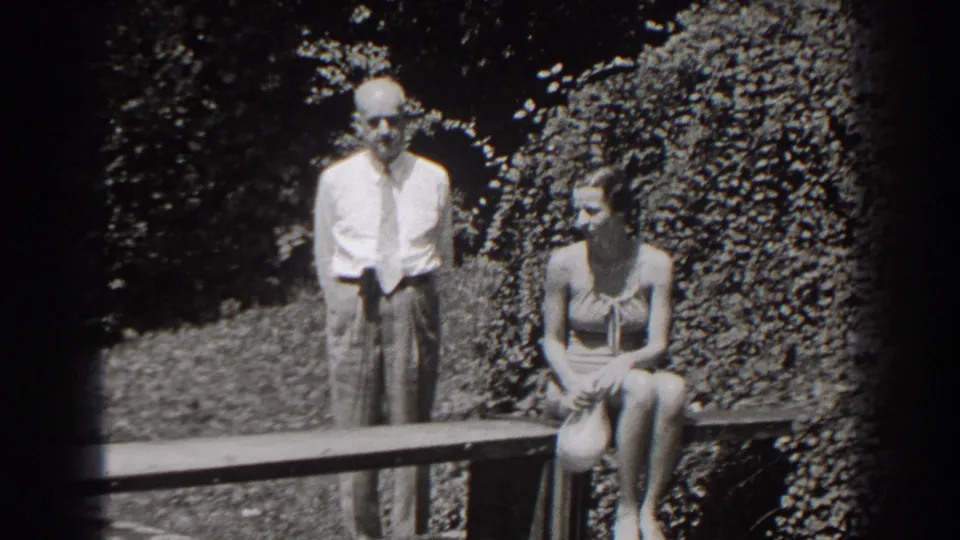 A 1938 New Jersey frame shows the woman seated on the bench and the older man standing behind the bench rail against dense foliage.