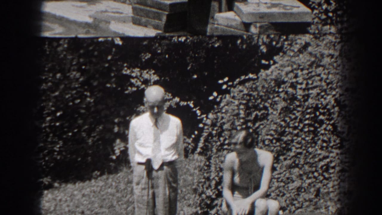 Man standing beside seated woman on a garden bench, with foliage and stone steps behind them.