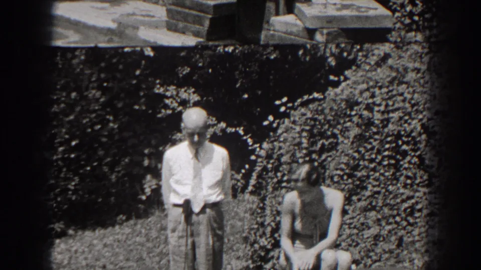 In 1938 New Jersey home-movie footage, a man in a light shirt stands near a woman seated on a wooden bench in a leafy garden.