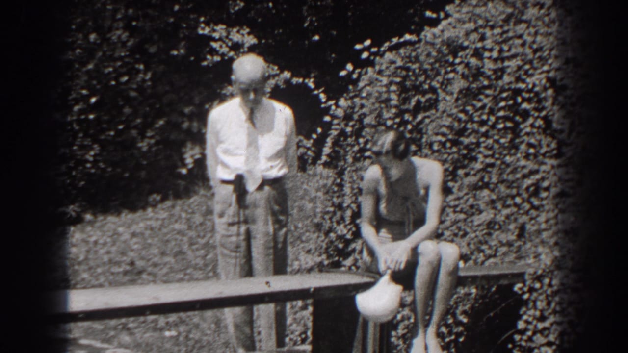 Man standing near a woman seated on a bench holding a light-colored cap in a garden.