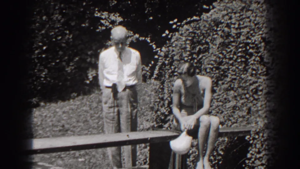 A man in a shirt and tie stands beside a seated woman holding a white cap, framed by garden foliage in New Jersey footage from 1938.