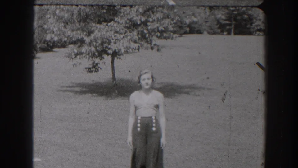 A young woman in a sleeveless top and high-waisted pants stands in a grassy New Jersey yard, with a small tree casting shade behind her.