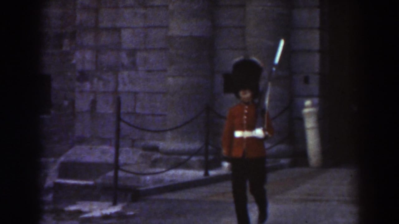 Red-coated guard with rifle marches in front of stone masonry and chain barriers.