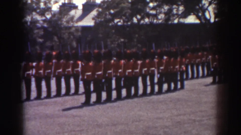 1938 Quebec home-movie frame showing uniformed men in red tunics standing in formation with rifles in a courtyard.