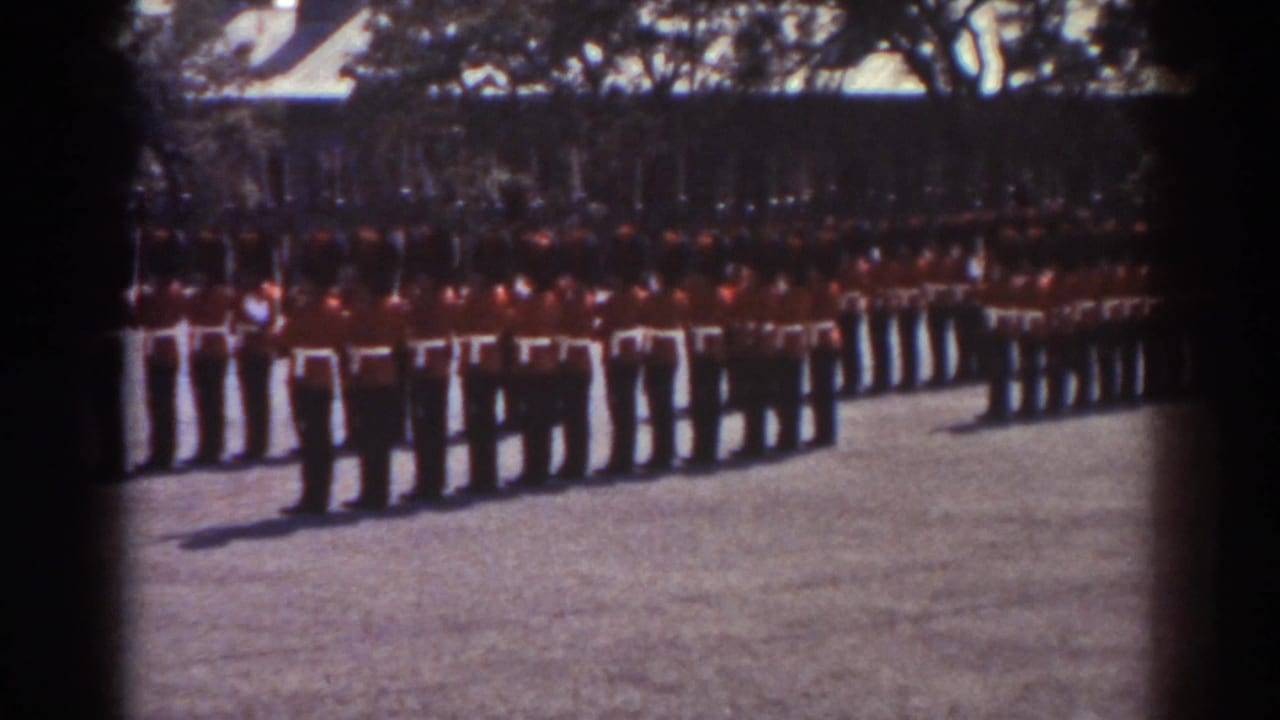 1938 Quebec home-movie frame showing a distant ceremonial formation of uniformed men with rifles in a paved open area.