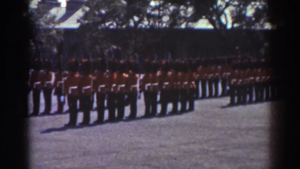 1938 Quebec home-movie frame showing a long formation of soldiers in red uniforms, with rifles and shaded trees in the background.