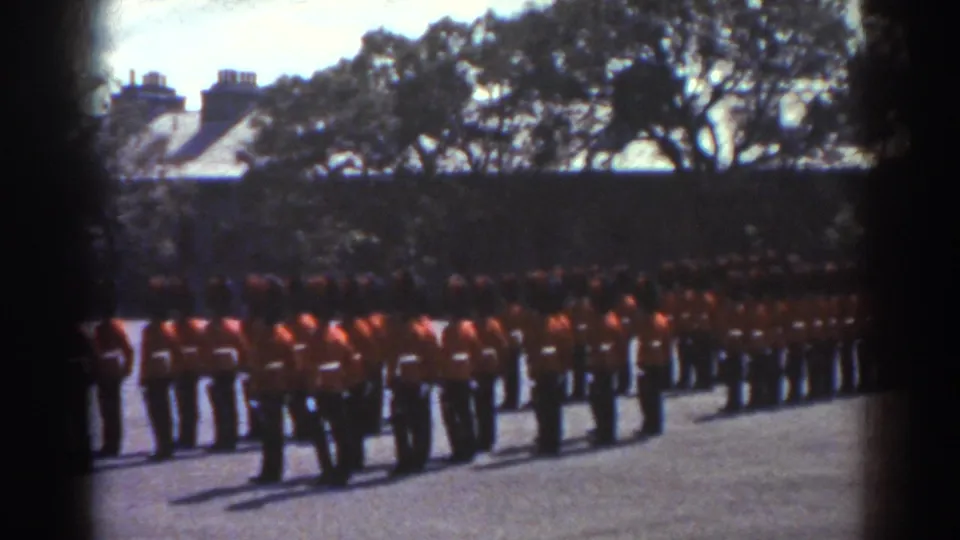 1938 Quebec frame showing soldiers in red tunics and dark hats standing in organized ranks on an outdoor parade ground.