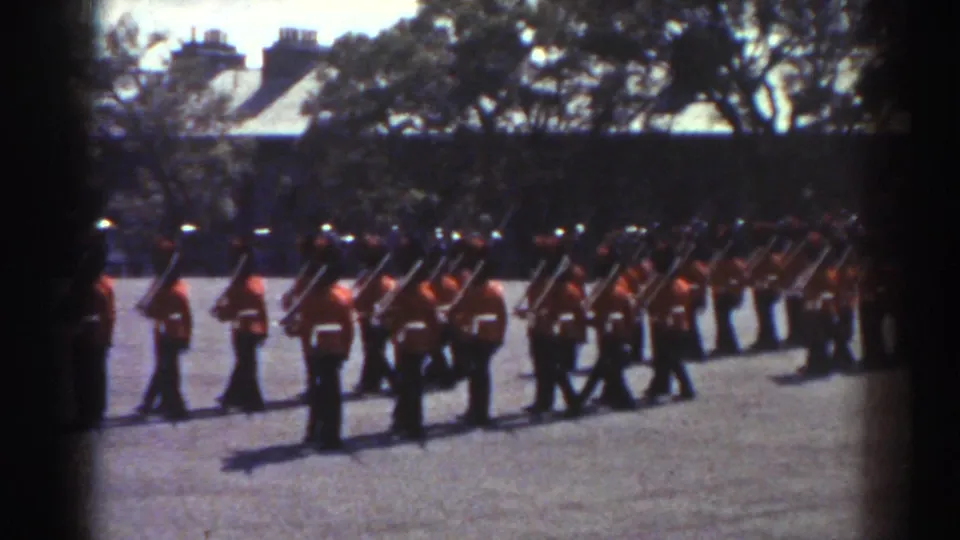 1938 Quebec frame of a ceremonial unit in red tunics and tall dark hats lined up with rifles outdoors.