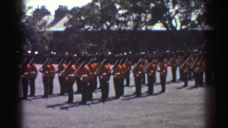 Wide 1938 Quebec home-movie view of red-uniformed ceremonial ranks with rifles in an outdoor drill area.