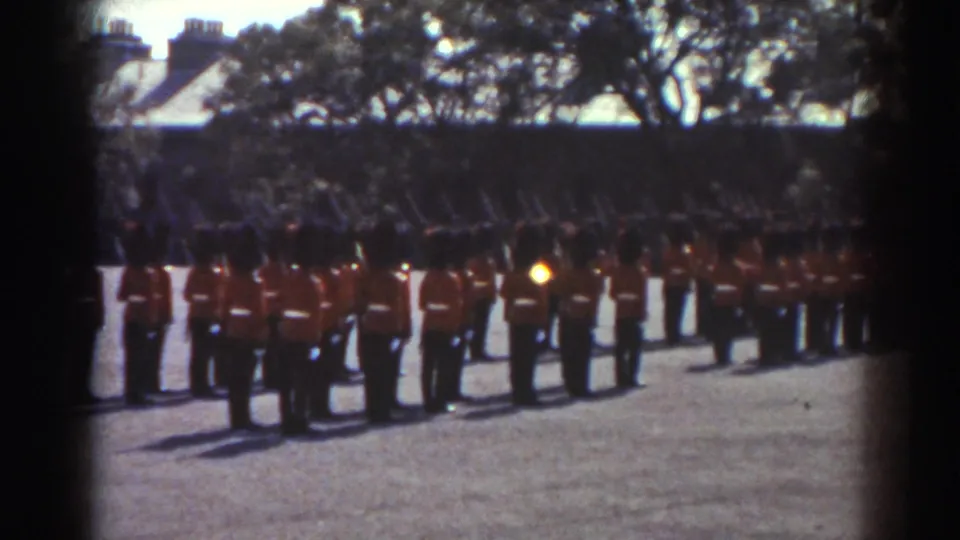 In Quebec, 1938, uniformed guards in red coats and tall dark hats stand in ranks during a drill on an open paved ground.