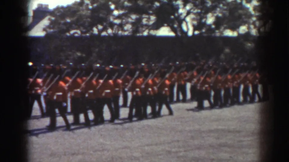 In the supplied 1938 Quebec context, guards in red tunics and dark hats move across the parade ground with rifles held in drill position.
