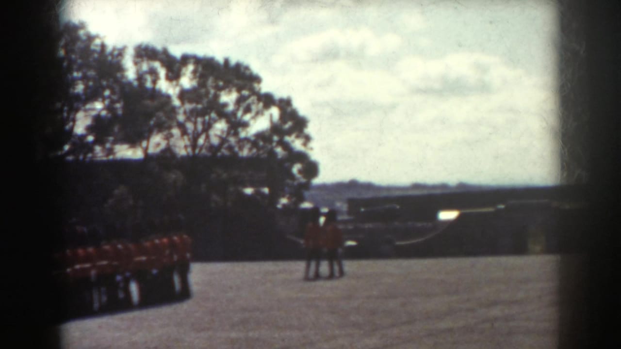 1938 Quebec home-movie frame showing red-uniformed guards along the left side of a bright paved courtyard with trees and distant structures.