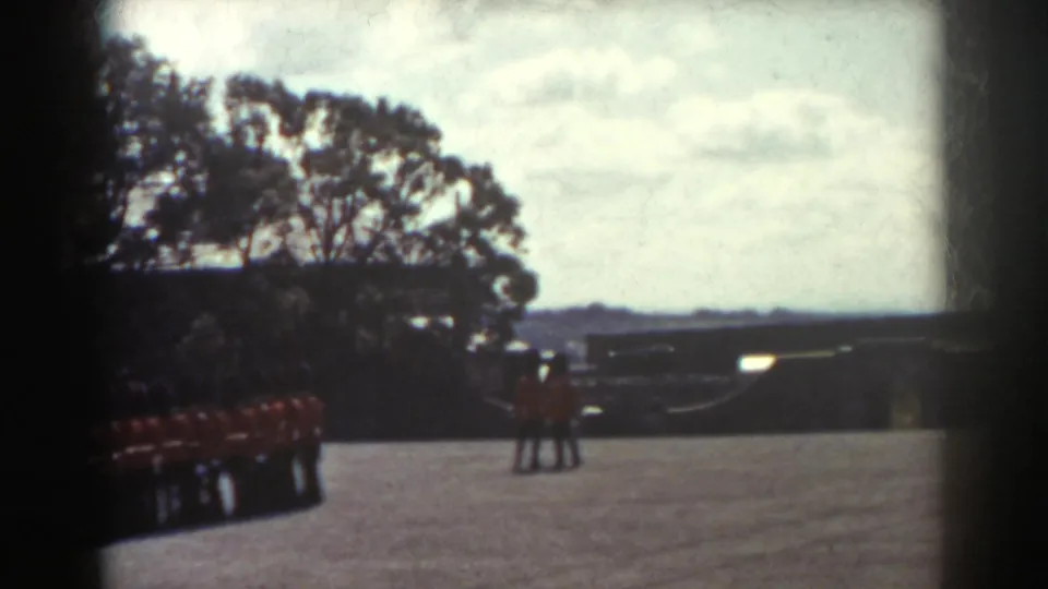 1938 Quebec home-movie frame showing red-uniformed guards along the left side of a bright paved courtyard with trees and distant structures.
