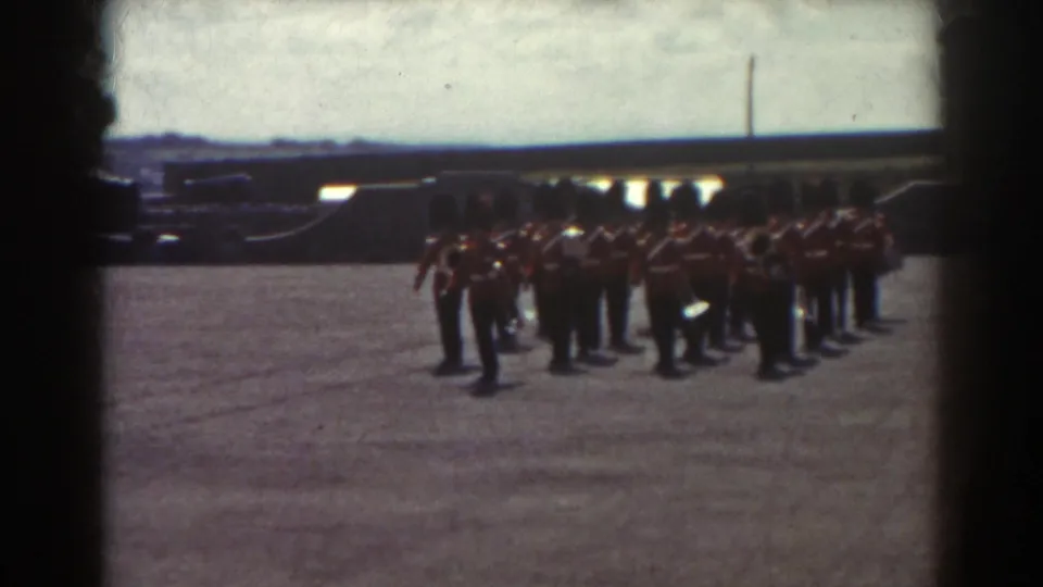 1938 Quebec frame showing red-uniformed guards in tall dark hats marching in formation across a sunlit paved courtyard.