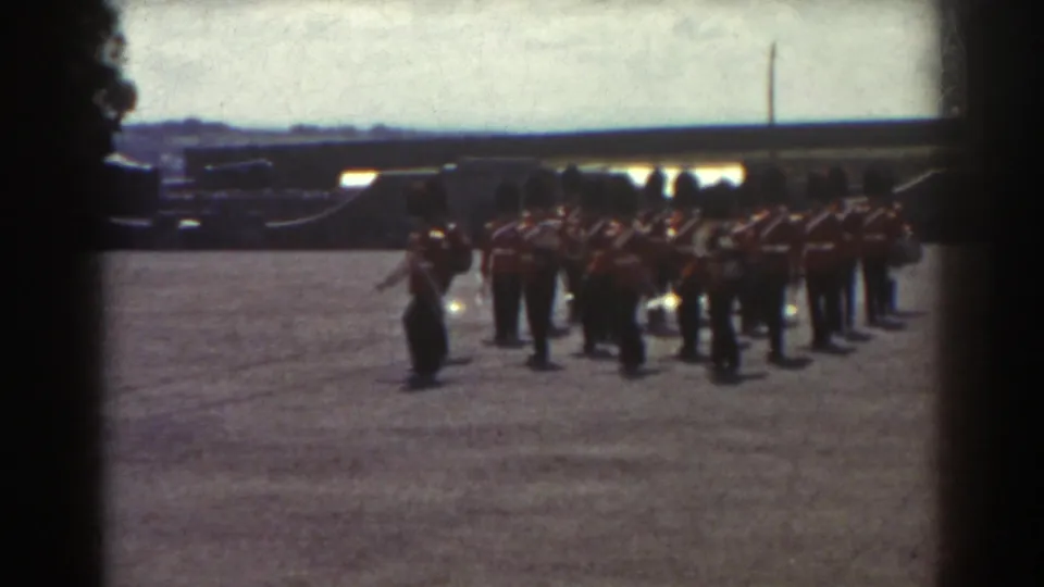 1938 Quebec home-movie frame showing red-uniformed guard band members forming a line on a bright gravel parade ground.