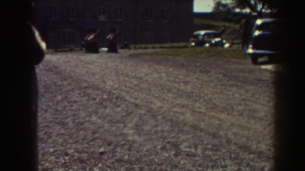 Quebec 1938 home-movie frame showing a gravel courtyard, period cars, and a stone building before the soldier formation appears clearly.