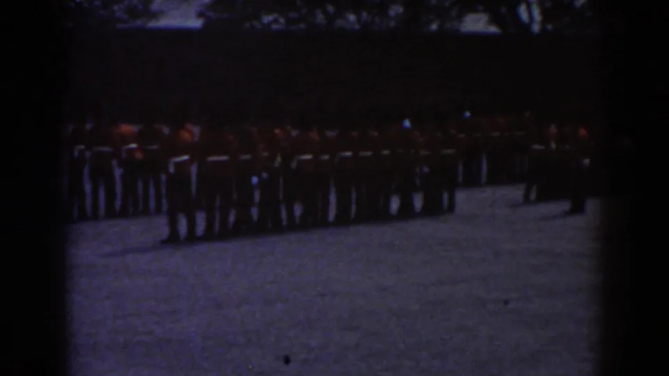 Quebec 1938 frame showing soldiers in red tunics and tall dark hats lined up in formation on a gravel courtyard.