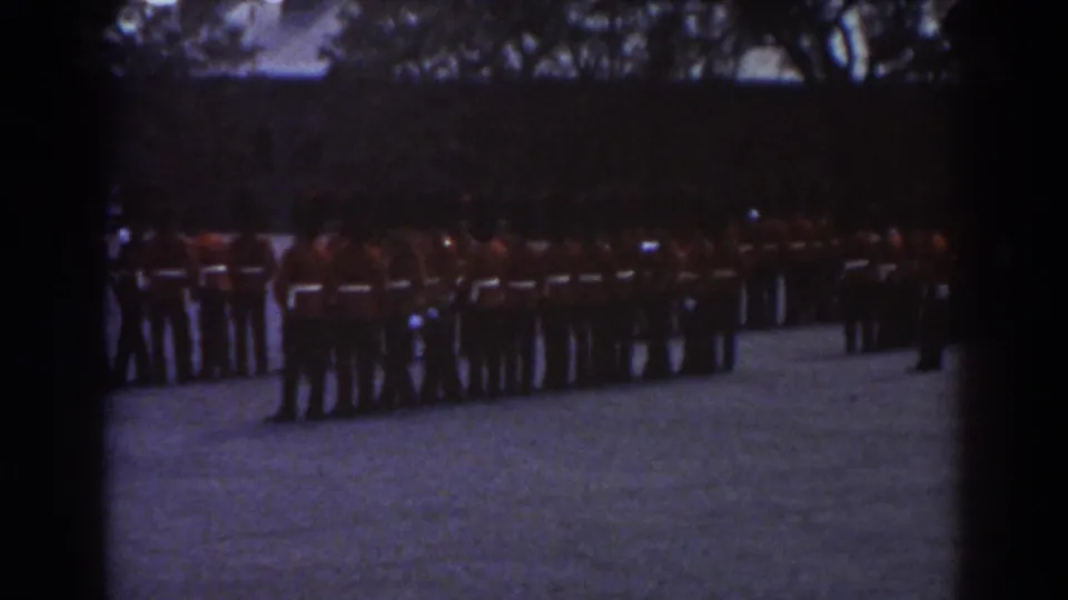Quebec 1938 home-movie frame with red-uniformed soldiers standing in formation across a shaded gravel courtyard.