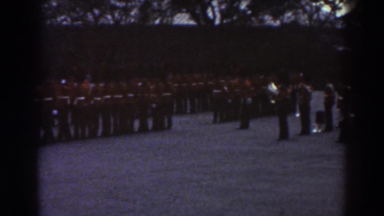 1938 Quebec home-movie frame showing uniformed soldiers lined up across a gravel field under dark tree cover.
