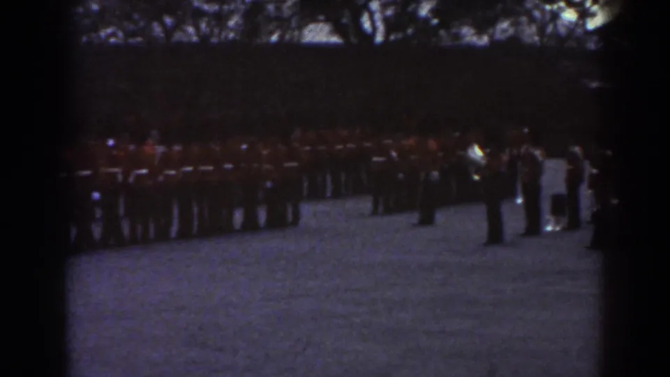 1938 Quebec home-movie frame showing uniformed soldiers lined up across a gravel field under dark tree cover.